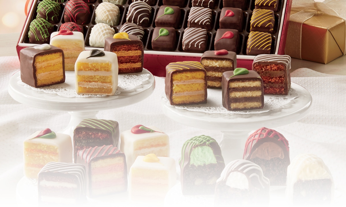 A boxed assortment of Bonbons and Petits Fours; a number of Petits Fours are arranged on plates and sliced to reveal layers of cake and buttercream.